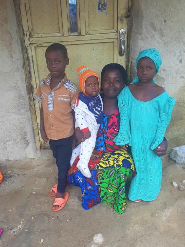 Saratu's children stand around her, as she kneels and smiles to the camera