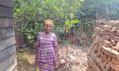 A little center-left, Vumilia, a Women for Women International participant, stands in a striped dress between two brick buildings