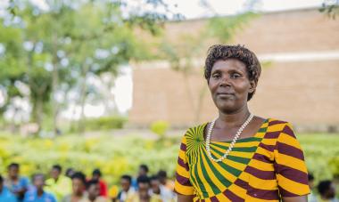 Caritas standing in a patterned shirt off to the right, looking proudly into the camera. Behind her is a class she trains at Women for Women International in Rwanda.