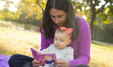 Kara, a woman, sitting outside in the grass, with a child in her lap and a book, reading to her